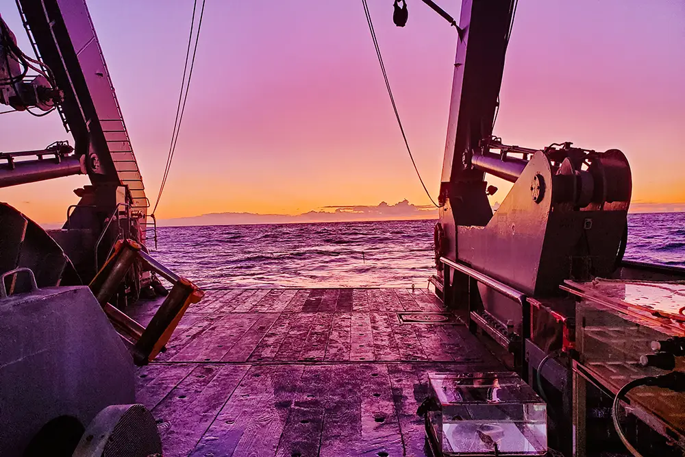 View from the deck of a research vessel at sunset, framed by large metal cranes and equipment, looking out over a calm ocean under a vibrant pink, purple, and orange sky.